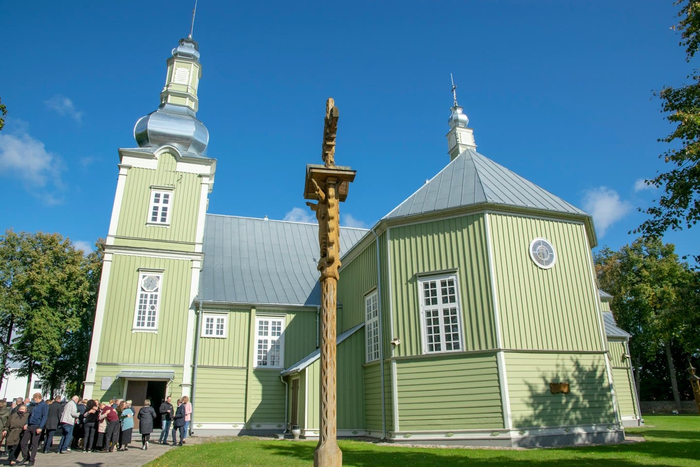 Persone riunite fuori da una chiesa in legno verde chiaro con un campanile.
